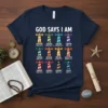 Navy blue t-shirt displaying "GOD SAYS I AM" above 12 colorful lighthouses, each paired with a descriptive word and Bible