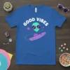 Good Vibes text above a cartoon alien wearing sunglasses, riding a UFO with planets and stars, on a royal blue