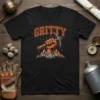 Black t-shirt with the word "GRITTY" in orange arching text above a fist holding a pickaxe with mountain graphics