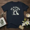 He Is Risen Easter T-Shirt with empty tomb graphic and doves, cream color text on navy shirt