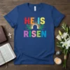 He Is Risen text design with colorful letters and a sunburst graphic, featured on a blue t-shirt