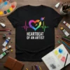 Black t-shirt with white text "Heartbeat of an Artist" above a rainbow heartbeat graphic with a paintbrush