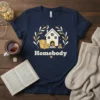 Homebody t-shirt with a cozy house illustration, armchair, and fireplace, featuring the word Homebody in bold text