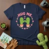 Hug Me Cactus T-Shirt with two smiling cacti in pots, surrounded by a floral wreath. Primary keyword: Hug Me Cactus