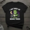 Frankenstein character sitting on a tombstone, holding coffee, with text "I Am Tired Of Being Tired" on a black t-shirt