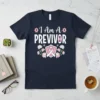 Navy t-shirt with white text "I Am A PREVIVOR" and pink ribbon graphic, surrounded by pink flowers and green leaves