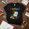 Black t-shirt with "i arted" text in rainbow colors, featuring an easel with a colorful paint splash and cartoonish paint