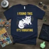 Navy blue t-shirt with yellow text "I FOUND THIS IT'S VIBRATING" and a white cat being held by purple hands