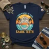 T-shirt design with text "I Judge Beaches By Their Shark Teeth" featuring a shark skull over a beach sunset scene