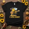 Cute bee character holding a honey dipper, dripping honey, with "I LOVE HONEY" text below on a black t-shirt