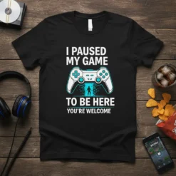 Black t-shirt with white text: "I PAUSED MY GAME TO BE HERE YOU'RE WELCOME", featuring a video game controller graphic