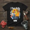 Black t-shirt with white text "I Turn Grills ON" and a cartoon man grilling over flames