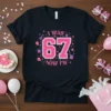 I WAS 67 NOW I'M text design in pink and white with birthday party graphics on a black t-shirt