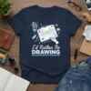 I'd Rather Be DRAWING t-shirt featuring a hand sketching a cityscape in an open notebook, surrounded by art supplies