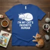 White text reading "I'M MY CAT'S FAVORITE HUMAN" with a sleeping cat graphic above it on a blue t-shirt