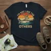 Vintage style t-shirt design with text "I'm Not Like The Others" above a deer and fish graphic with sunset background