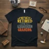 Black t-shirt with "I'm Not Retired I'm A Professional Grandpa" text in yellow, grey, and orange