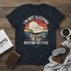 T-shirt design with text "I'm Not Sleeping I'm Just Resting My Eyes" featuring a vintage armchair on books and a