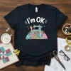 I'm OK t-shirt with a cartoon sewing machine, colorful thread spools, and a waving hand, set against a patterned background