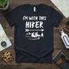 White text "I'm With This Hiker" on a navy t-shirt, with graphics of hiking boots, mountains, and trees