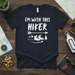 White text "I'm With This Hiker" on a navy t-shirt, with graphics of hiking boots, mountains, and trees