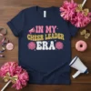 Navy blue t-shirt with "IN MY CHEER LEADER ERA" text in pink, gold glitter, and white retro font, featuring pink