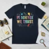 Navy blue t-shirt with colorful "In Science We Trust" text and science-themed doodles like DNA, lightbulb, and rocket