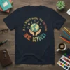 In A World Where You Anything Be Kind t-shirt featuring a globe held by hands and educational icons