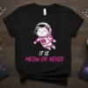 Black t-shirt with a cute cartoon cat astronaut floating in space, text reads "IT IS MEOW OR NEVER" in pink