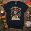 Skeleton playing guitar with "It's Día de los That's Why" text, floral, and butterfly accents on a black t-shirt