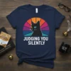 Navy t-shirt with "Judging You Silently" text and a black cat graphic looking sideways, featuring a retro sunset background