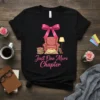Just One More Chapter quote on a black t-shirt with a graphic of a comfy armchair, books, and a lamp
