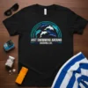 Black t-shirt with "Just Swimming Around Enjoying Life" text and a graphic of dolphins swimming under a rainbow arc
