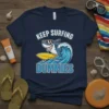 Keep Surfing Dummies t-shirt with a cartoon shark surfing a wave. The design features bold text and a fun, cartoony