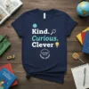Navy t-shirt with "Kind. Curious. Clever" in white and teal text, featuring a smiley face, magnifying glass, and lightbulb graphic