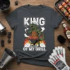 King Of My Grill T-Shirt featuring a T-Rex dinosaur wearing a chef hat and apron, grilling sausages on a flaming