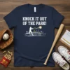 Baseball t-shirt with "Knock It Out Of The Park!" text, a batter hitting a ball, and stadium graphic