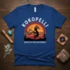 Kokopelli playing flute graphic on a blue t-shirt, text "Kokopelli Spirit of the Southwest", sunset and desert landscape design