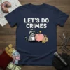 Let's Do Crimes t-shirt design with a raccoon putting coins into a piggy bank, white text on navy blue shirt