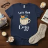Lets Get Cozy t-shirt design with an orange cat in a cable-knit coffee mug, wearing a blue scarf