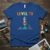 Level 73 text with a rocket launching over mountains and city skyline on a blue t-shirt