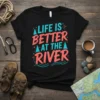 Black t-shirt with "Life Is Better At The River" text in coral and teal, featuring pine trees and wavy lines