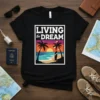 Black t-shirt with "LIVING the DREAM" text above a colorful beach sunset with palm trees and a surfboard, representing vacation