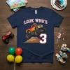 LOOK WHO'S 3 text with a red and yellow monster truck graphic on a navy blue t-shirt for a boy's