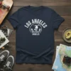 Los Angeles Hollywood Angeles text with a skateboarder graphic and palm trees, white design on navy t-shirt