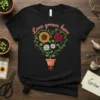 Black t-shirt with "Love Grows Here" text above a floral heart containing a sunflower, rose, and daisies in a pot