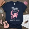 Navy t-shirt with white text "Taylor" and "MAGIC IS IN YOU", featuring a pink unicorn playing a guitar with musical