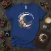 Magical Moon Blooms text on a t-shirt with a floral crescent moon graphic, featuring white and yellow flowers and greenery