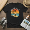 Black t-shirt with "Malibu Beach" text above a circular graphic of palm trees, a surfer, and a sunset over the