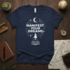 Navy blue t-shirt with "MANIFEST YOUR DREAMS" text, an open book, rocket, moon, and stars graphic